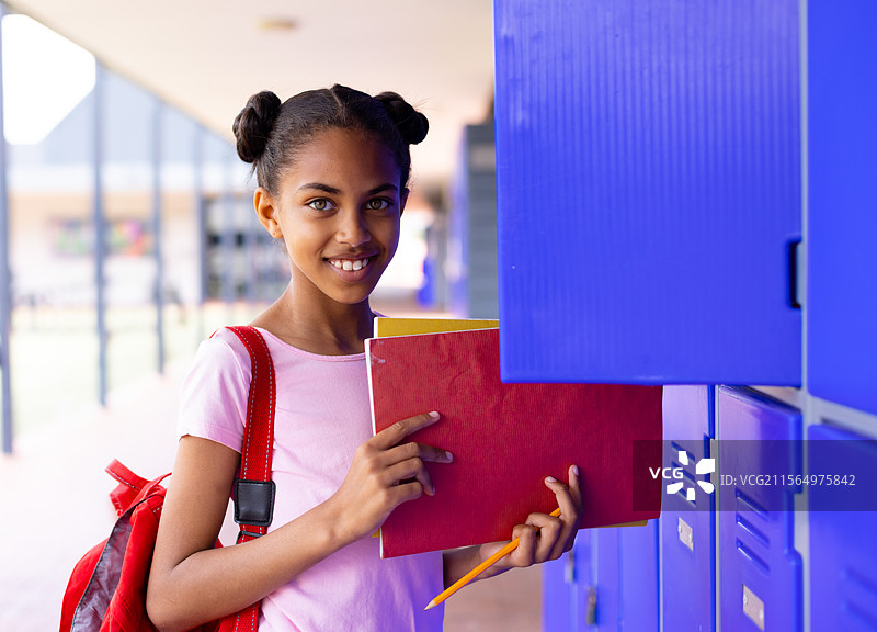 非裔美国少女在学校走廊使用储物柜，手里拿着红色文件夹和红色背包。图片素材
