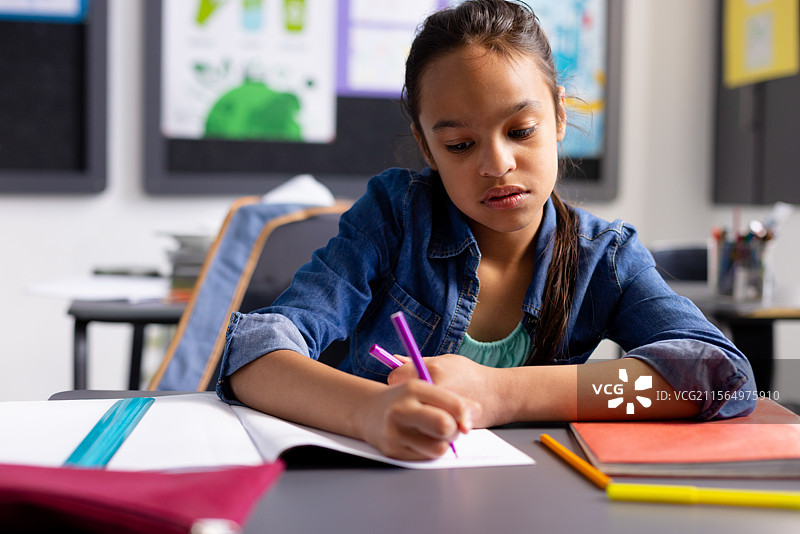 小女孩在小学教室的桌子上用紫色钢笔在笔记本上写字，旁边有一个活页夹，留有空白空间。图片素材