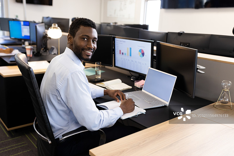 年轻的非裔美国男性在办公室桌子上微笑着看向镜头，手里写着笔记本，旁边有一台笔记本电脑。图片素材