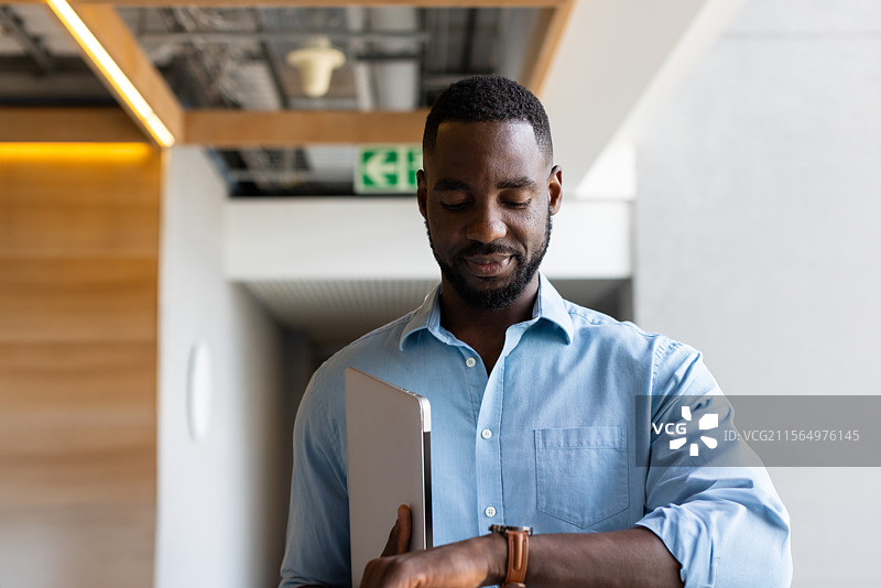 非裔美国男性在办公室走廊查看手表，同时手持笔记本电脑，旁边有出口标志图片素材