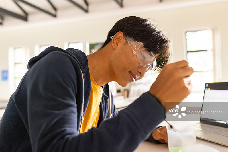 亚洲少年在科学实验室的桌子上用滴管吸取绿色溶液，旁边有笔记本电脑和护目镜。图片素材