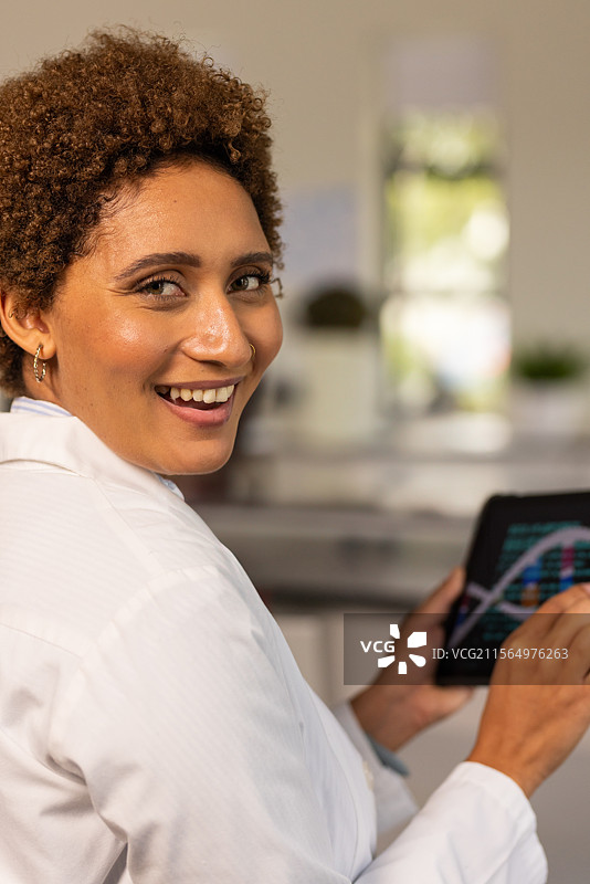 穿着白色实验室外套的女性在厨房实验室空间中转向镜头，手持带有图表的平板电脑。图片素材