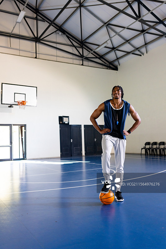 一位非裔美国男性站在室内篮球场上，脚踩篮球，佩戴哨子，留有空白空间。图片素材