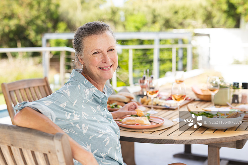 一位年长女性在户外的木质露台桌上享用午餐，桌上有三明治、沙拉碗和玫瑰红葡萄酒。图片素材