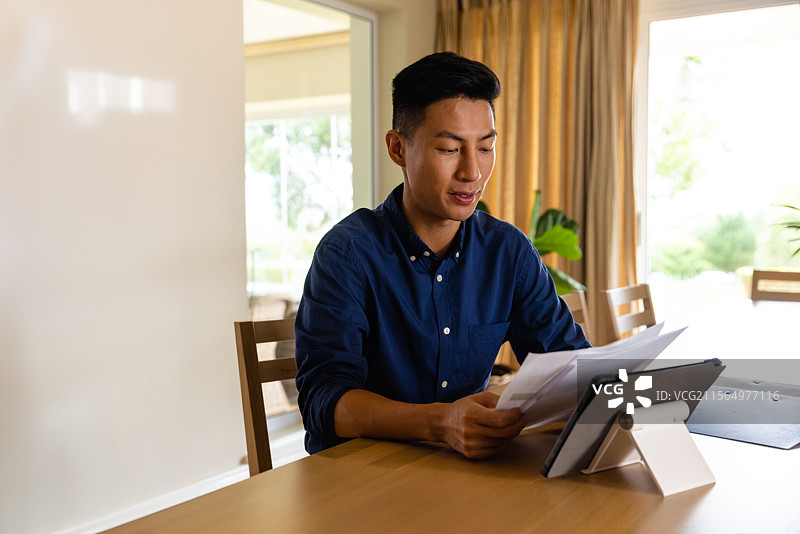 年轻的亚洲男子在家中的书房里，坐在木桌旁审阅文件，桌上有平板电脑和一盆植物。图片素材