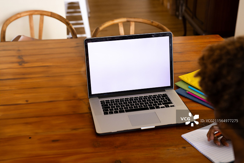 非裔美国人在家中的木质餐桌上写笔记，旁边有笔记本电脑和文件夹。图片素材