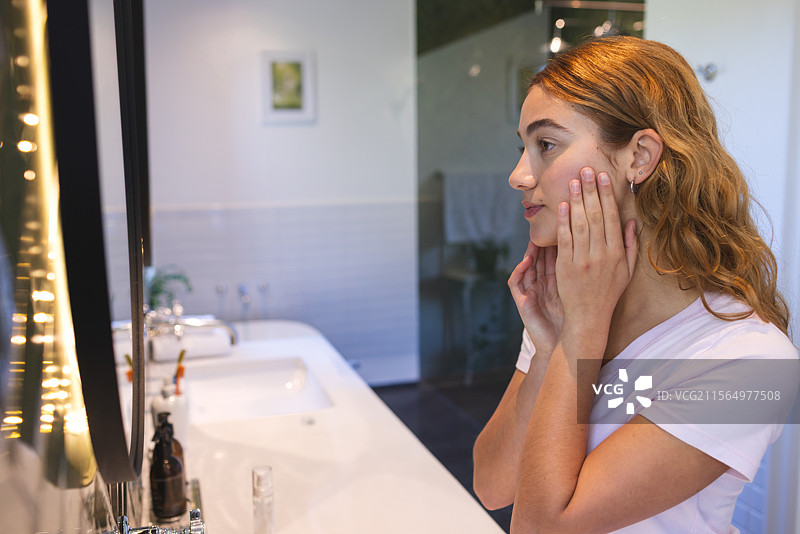 女人站在浴室的照明镜子前，轻触脸颊，周围有洗漱用品，留有空白空间。图片素材