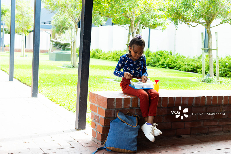 小女孩在院子里的砖墙上吃午餐，旁边有托盘、瓶子和背包，留有空白空间。图片素材