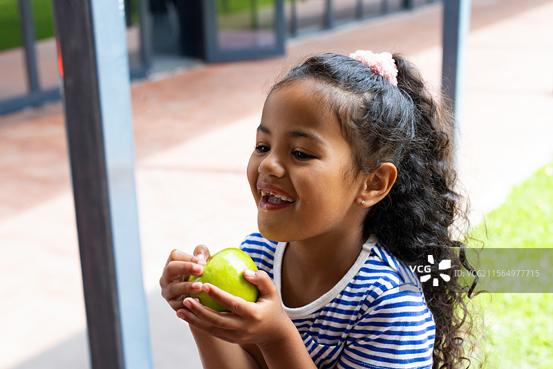 多种族女性儿童坐在瓷砖露台上，手里拿着一个绿色苹果，靠近玻璃门入口。图片素材