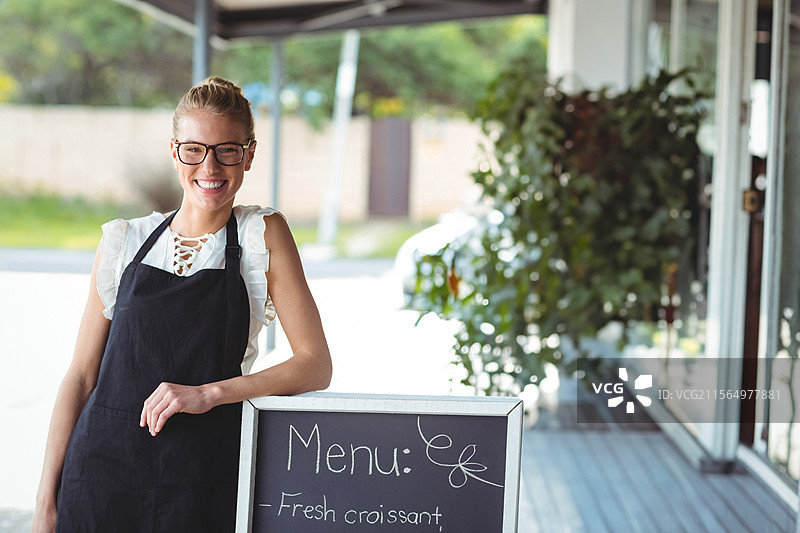 穿着围裙和眼镜的女性微笑着倚靠在咖啡馆遮阳篷下的黑板菜单标志上。图片素材