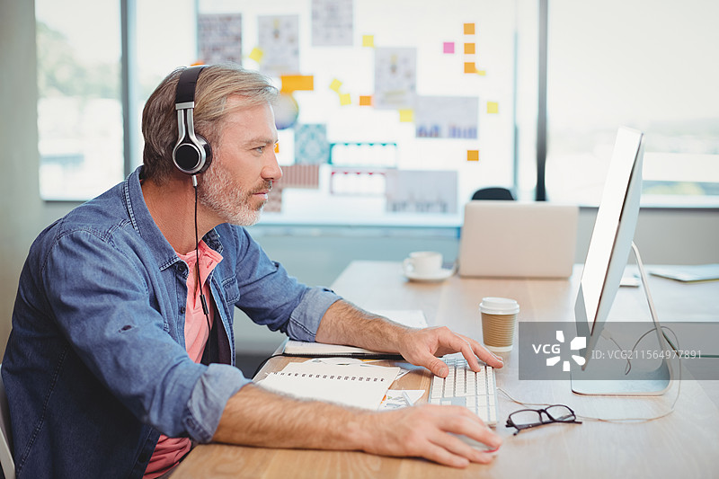 中年男子在窗边的办公室里使用白色键盘打字，佩戴耳机，旁边有咖啡杯，留有空白空间。图片素材