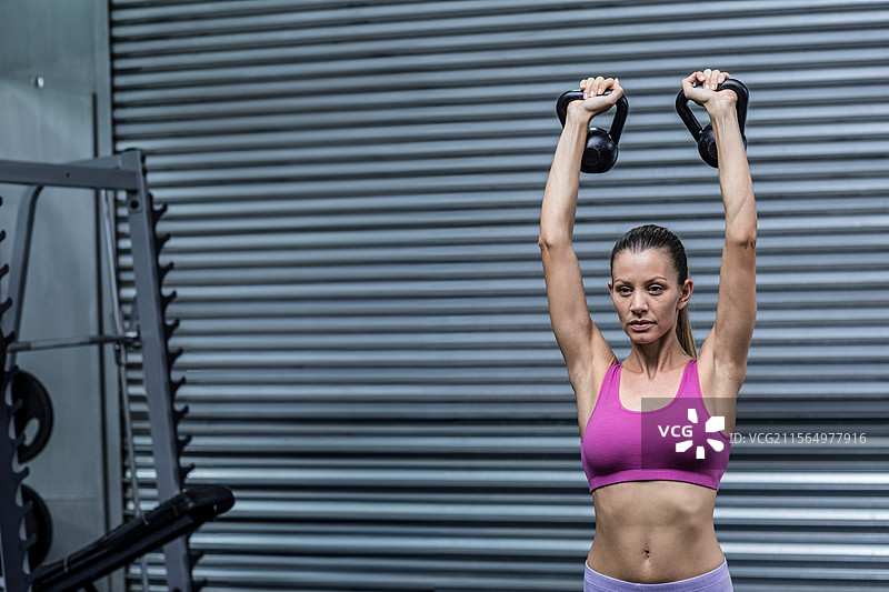 女性在健身房中举起壶铃，背景有深蹲架和装载的重物，留有空白空间图片素材
