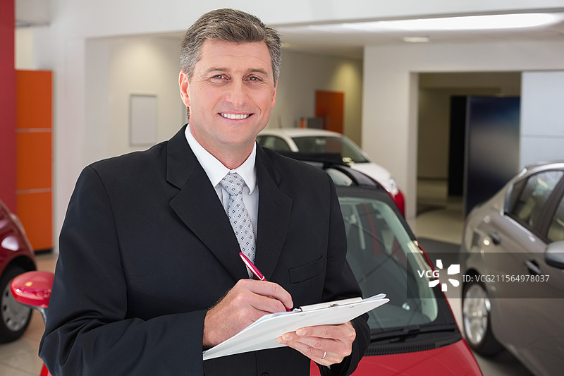 男子站在汽车经销商展厅内，手持夹板和红色圆珠笔，周围是新车。图片素材