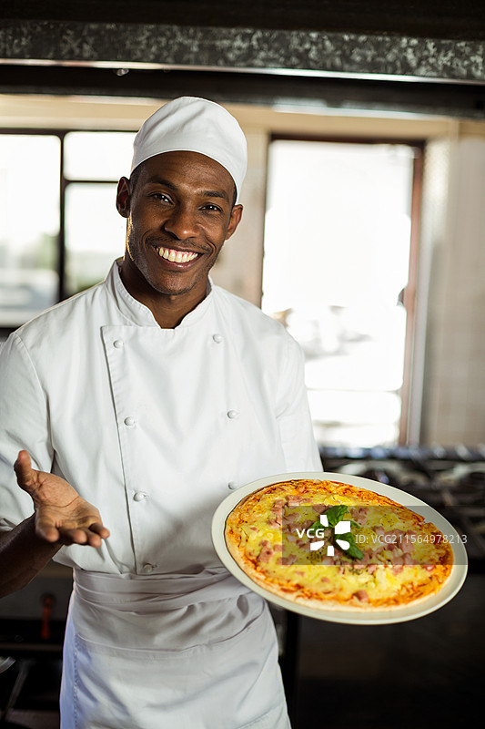 非裔美国男性厨师微笑着在商业厨房中展示新鲜出炉的比萨饼图片素材