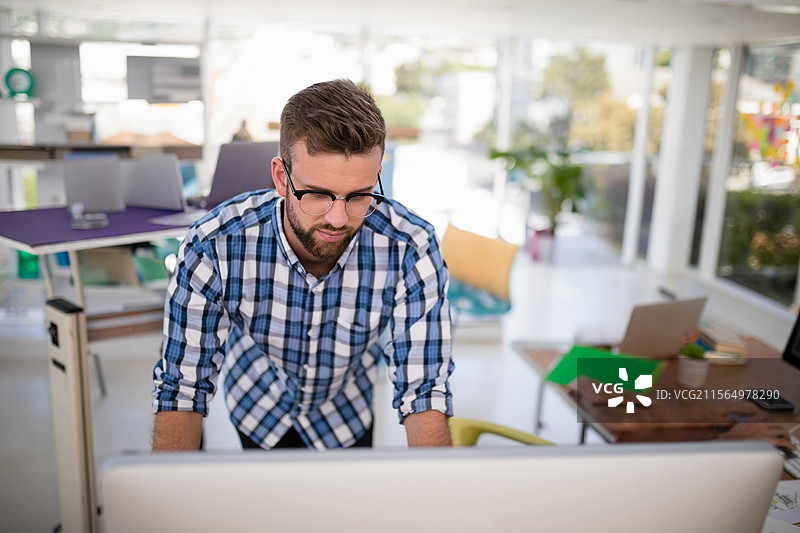 男子在现代开放式办公室中倾身靠近大型桌面显示器，旁边有笔记本电脑和智能手机。图片素材