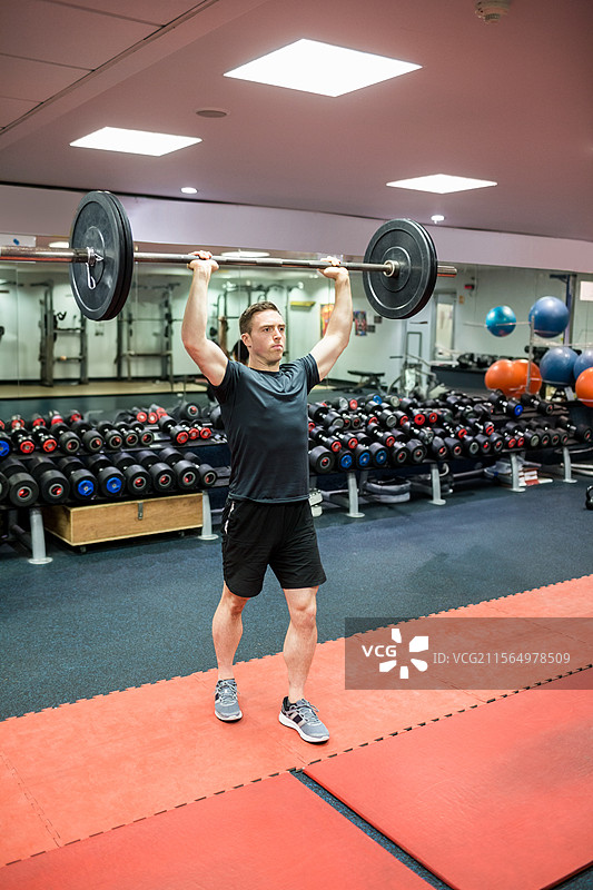 在健身房举起杠铃的男子，周围有哑铃架和垫子。图片素材