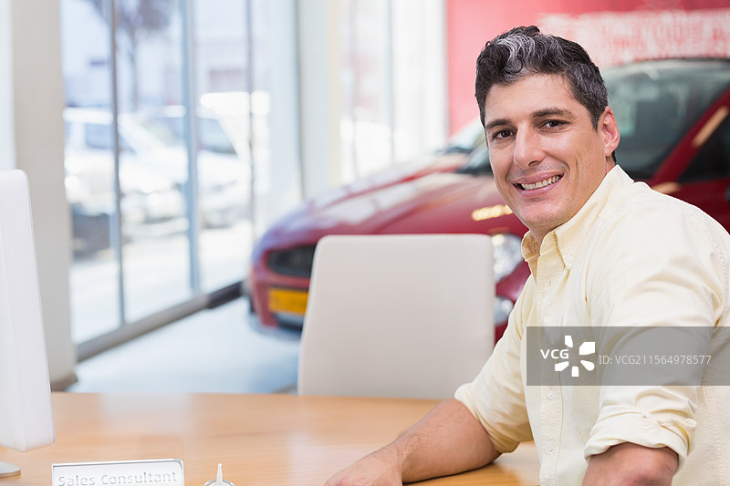 微笑的男子坐在明亮的汽车展厅内的木桌后，旁边是一辆红色展示车，留有空白空间。图片素材