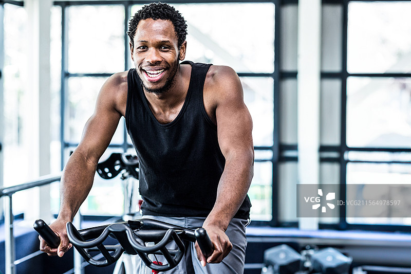 在现代健身房里，一位非裔美国男性正在健身车上骑行，同时手握哑铃。图片素材