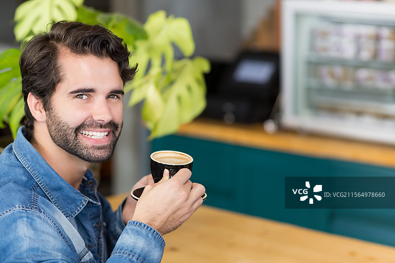 微笑的男人在咖啡馆桌子旁拿着黑色陶瓷咖啡杯，旁边有玻璃糕点展示柜，留有空白空间图片素材