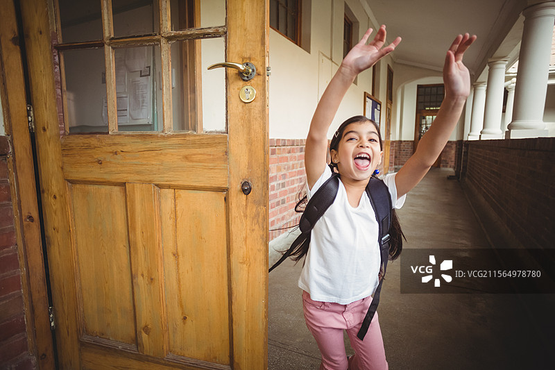 在学校走廊中，举起双臂的女童，身旁有木门和背包，留白空间。图片素材