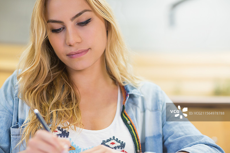 穿着牛仔衬衫的女性靠在书房的桌子上，手里拿着笔和笔记本。图片素材