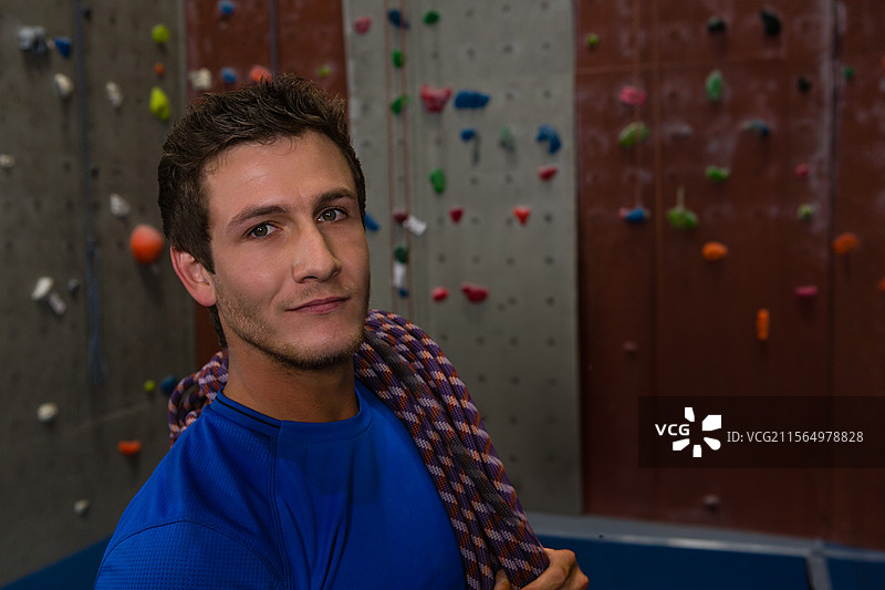 站着的男子将攀岩绳搭在肩上，在攀岩馆内，背景有五彩缤纷的攀岩点，留有空白空间。图片素材
