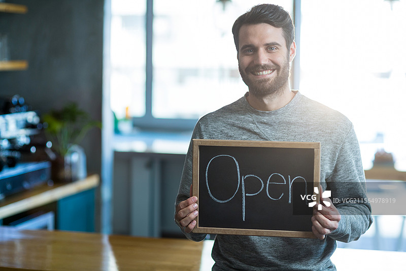 微笑的男子在咖啡馆柜台后面拿着写有“开放”的木质黑板招牌，旁边有一台咖啡机。图片素材