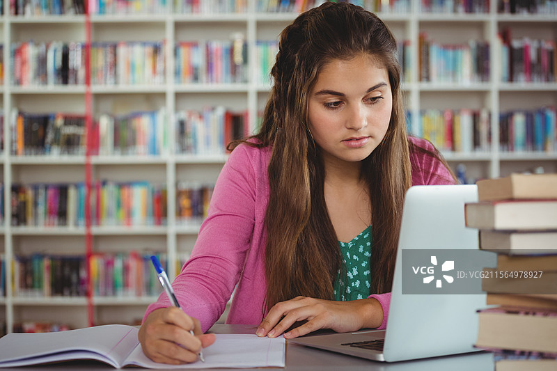 在图书馆桌子上学习的少女，正在用蓝色墨水的钢笔在笔记本上写字，旁边有一台银色的笔记本电脑。图片素材