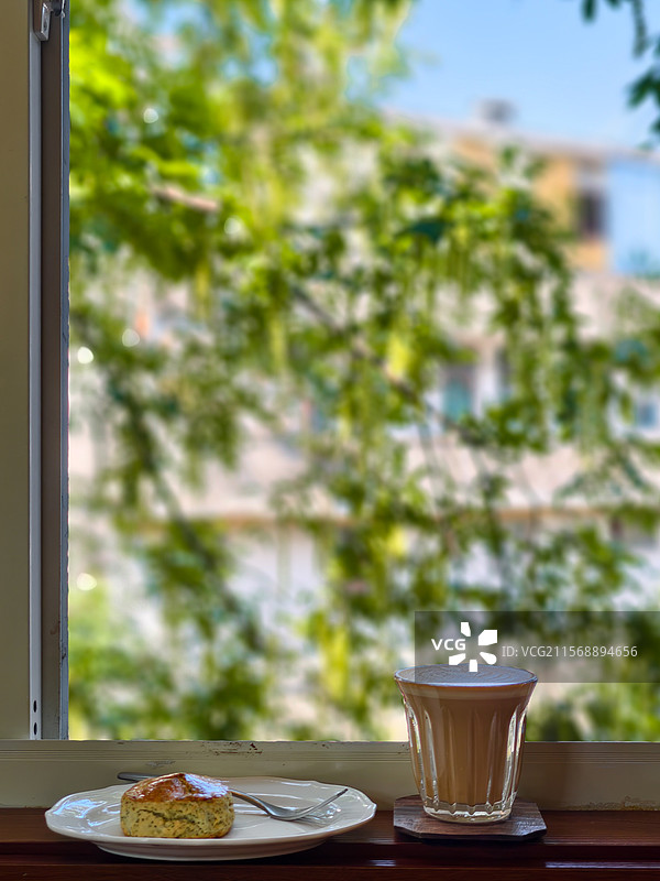 咖啡店玻璃窗外是被光打亮的绿树窗内窗台上是一盘司康和一杯澳白咖啡图片素材