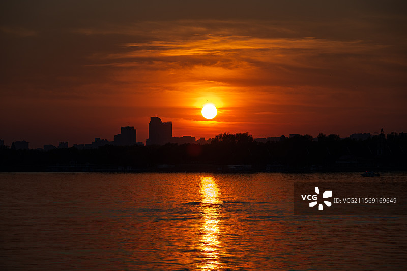 黑龙江哈尔滨松花江畔浪漫的日落晚霞，橘色天空与城市建筑天际线剪影，红日在江面形成倒影图片素材