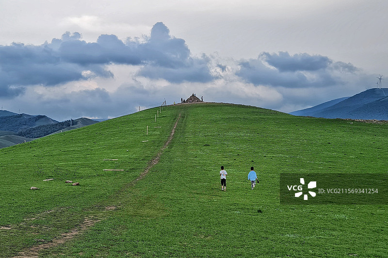 广阔草原上，两个孩子在山坡上行走，山顶有游客聚集，背景是多云的天空和远处的风力发电机。图片素材