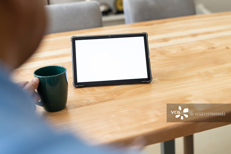 男人坐在家里的木桌旁，手握绿色杯子，正在查看平板电脑屏幕。图片素材