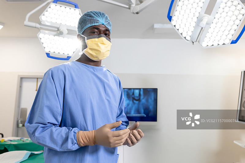 非裔美国男性外科医生在手术室内戴上手套和口罩，周围是手术灯。图片素材