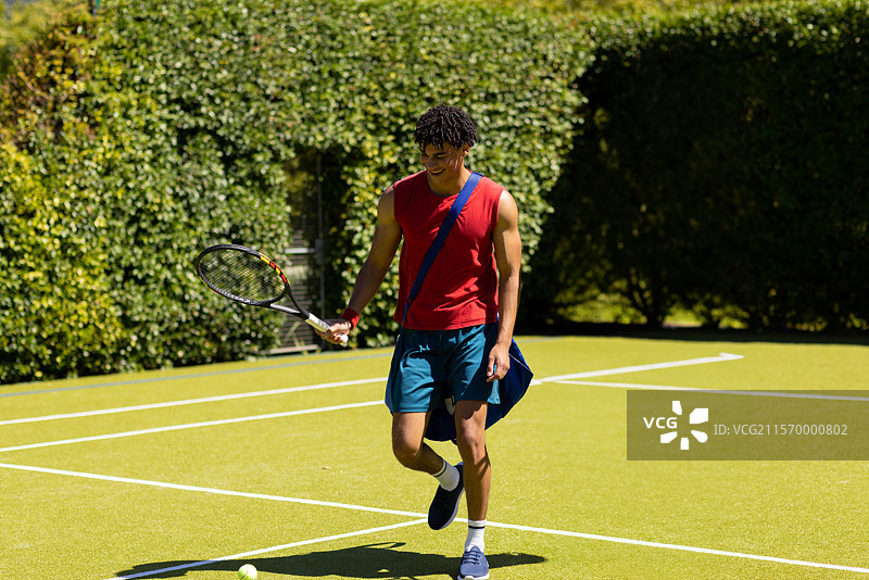 年轻男子在网球场上走，手里提着球拍和装有网球的蓝色运动包图片素材