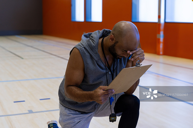 中年男性教练跪在室内篮球场上，用夹板和秒表擦汗图片素材