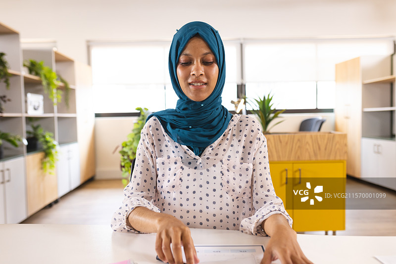 年轻女性在现代办公桌前审阅文件，桌上有植物和黄色柜子。图片素材