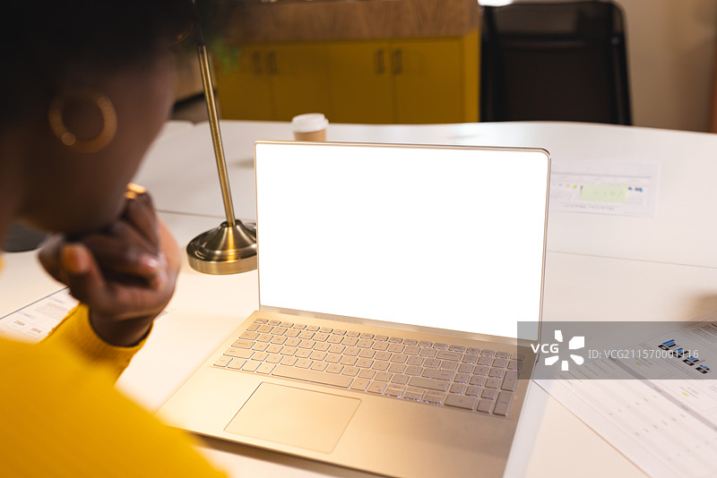 非裔美国女性在办公室的桌子上工作，查看发光的笔记本电脑和黄铜台灯。图片素材
