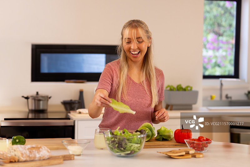 年轻女性在现代厨房的岛台上准备玻璃碗里的沙拉，旁边有面包和西红柿。图片素材