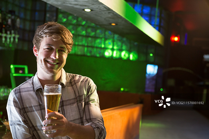 微笑的男人站在昏暗的酒吧吧台前，手里拿着高脚啤酒杯，留有空白空间图片素材