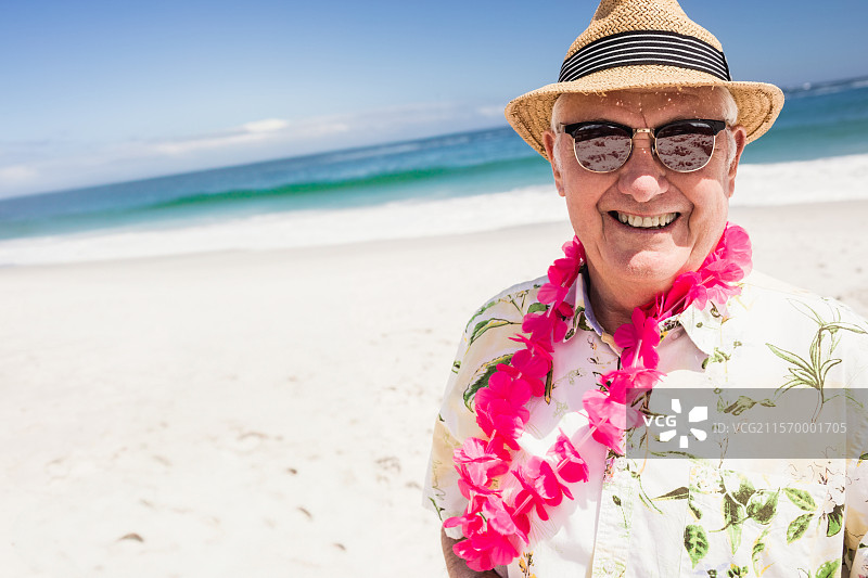 戴着草帽、太阳镜和花环的老年男子站在晴朗天空下的沙滩上图片素材