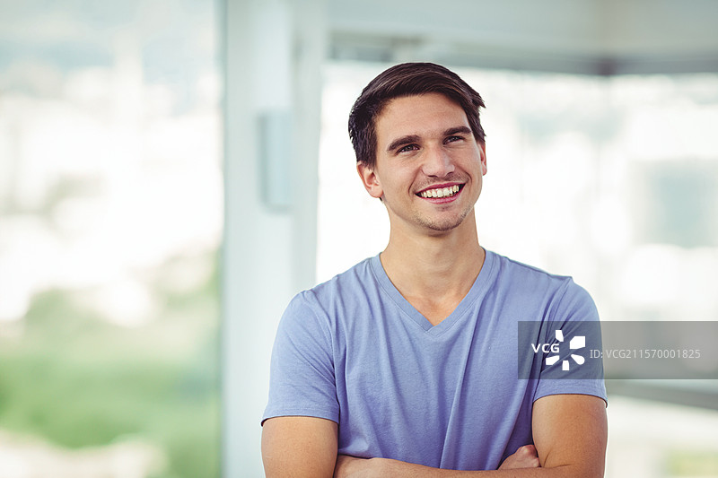 微笑的男性，二十多岁，双臂交叉站在现代室内，背景是模糊的城市景观。图片素材