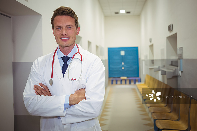 微笑的男性医生在医院走廊交叉双臂，穿着白大褂，佩戴红色听诊器，留有空白空间图片素材