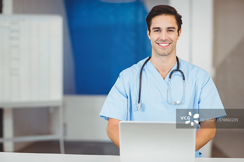 微笑的男护士坐在诊所桌子旁，手边有笔记本电脑、听诊器和医疗记录，留有空白空间。图片素材