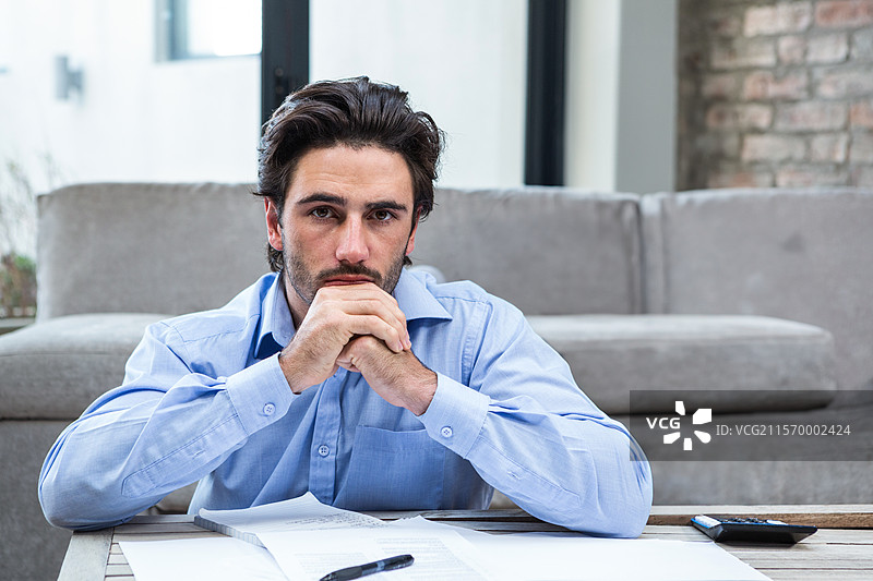 靠在桌子上的男人用手托着下巴，周围是散落的文件和计算器，坐在客厅里。图片素材