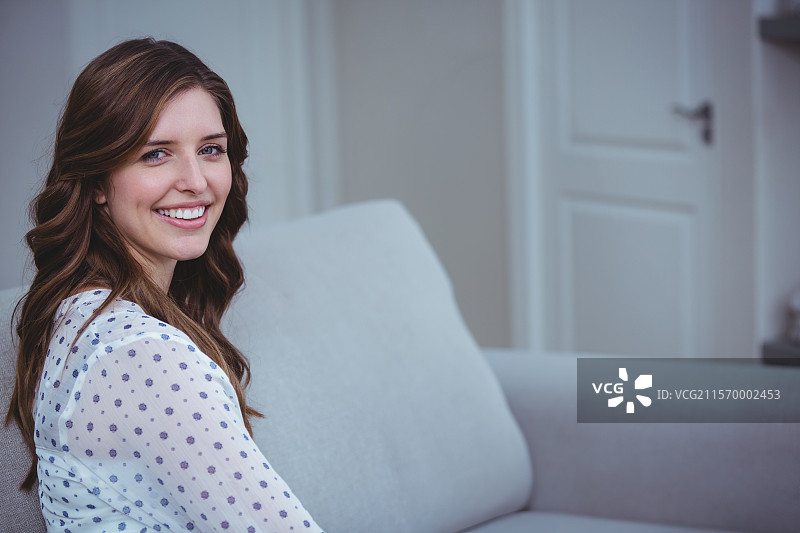 微笑的女性坐在浅灰色的软垫沙发上，突显出简约的室内装饰。图片素材