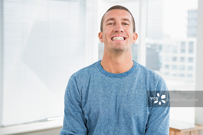 微笑的男子穿着蓝色毛衣，站在现代办公室工作室里，窗帘和城市天际线尽收眼底。图片素材