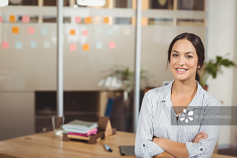 微笑的女性在办公室里双臂交叉，身边有笔记本和便利贴，留有空白空间。图片素材