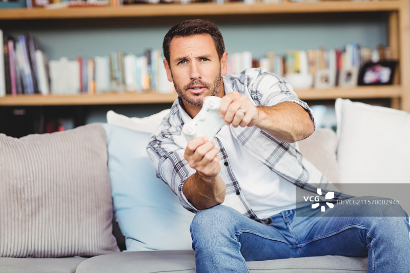 紧握白色游戏手柄的男子，向前倾身坐在客厅沙发上，身后是一个木制书架。图片素材