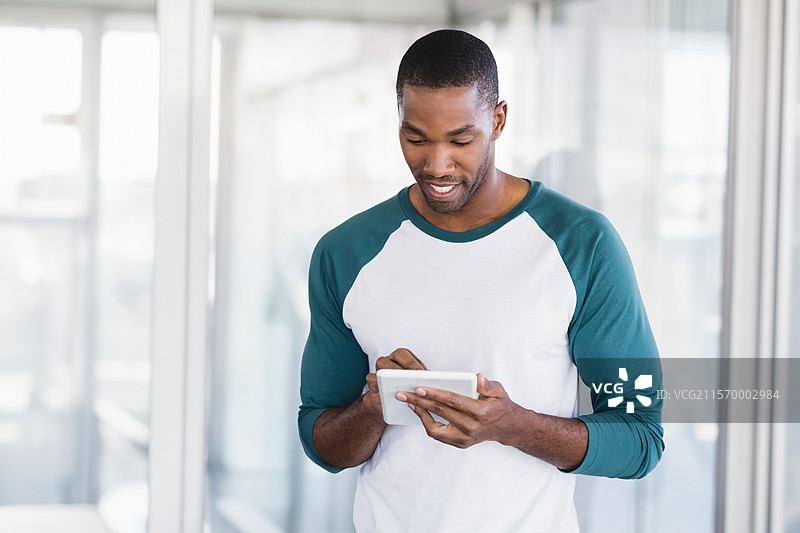 在现代办公室走廊中，一位中年非裔美国男性正在使用平板电脑，周围有玻璃隔断。图片素材