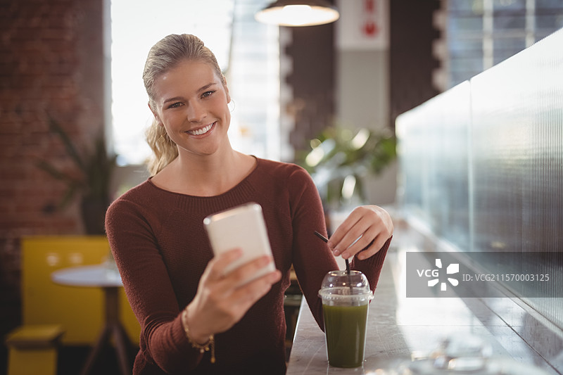 在咖啡馆柜台前，女性正在用智能手机拍自拍，搅拌果昔，旁边有塑料杯，留有空白空间。图片素材
